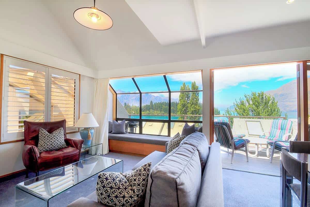 A modern living room with a glass coffee table, a sofa, and a chair. There are large windows offering a view of the outdoors.