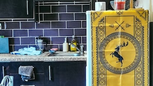 Eine Küche mit dunklen Schränken, einer gefliesten Spritzschutzwand und einem gelben dekorativen Banner mit einem Hirsch-Emblem und dem Text 'OURS IS THE FURY'.