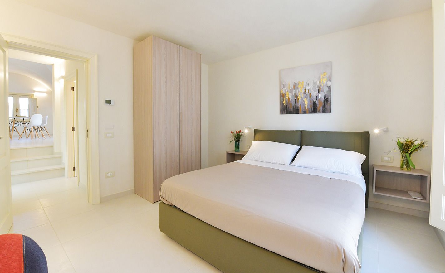 A modern bedroom with a large bed, a wardrobe, a painting on the wall, and a nightstand with a vase.