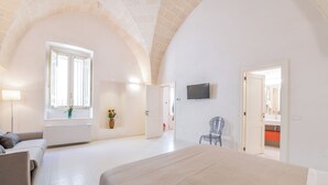 A spacious bedroom with a large bed, a TV mounted on the wall, and a window with white shutters. - Casa Magadè (Manduria)