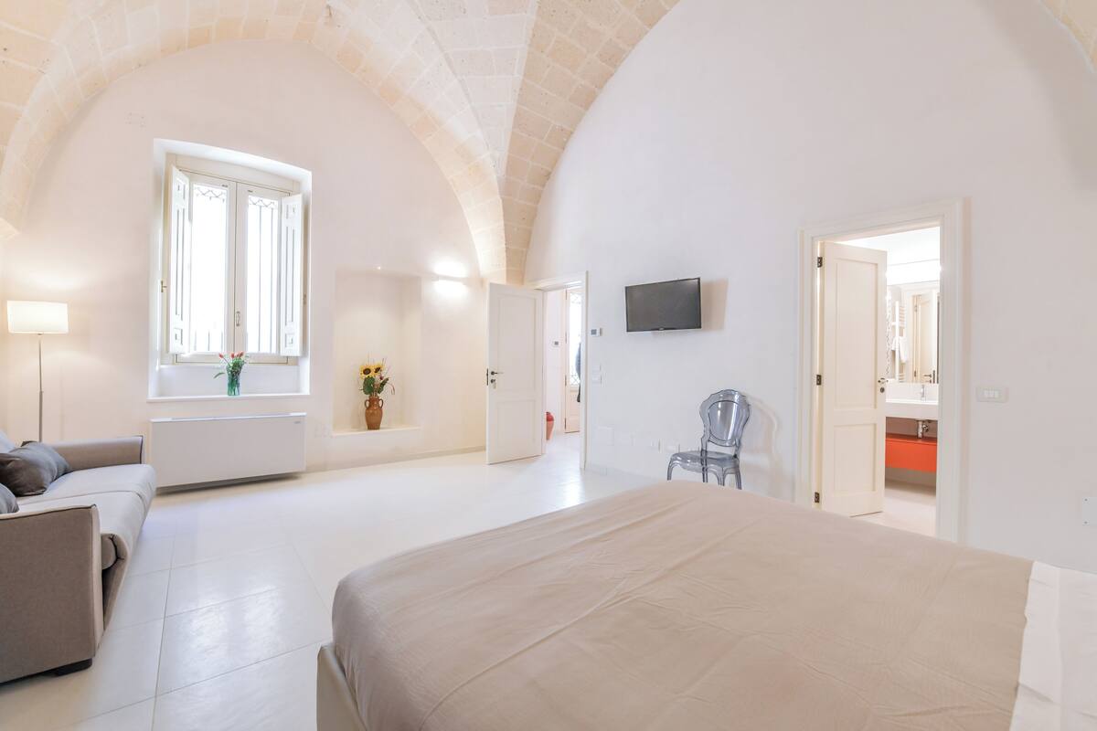 Un dormitorio espacioso con una cama grande, un televisor montado en la pared y una ventana con contraventanas blancas.