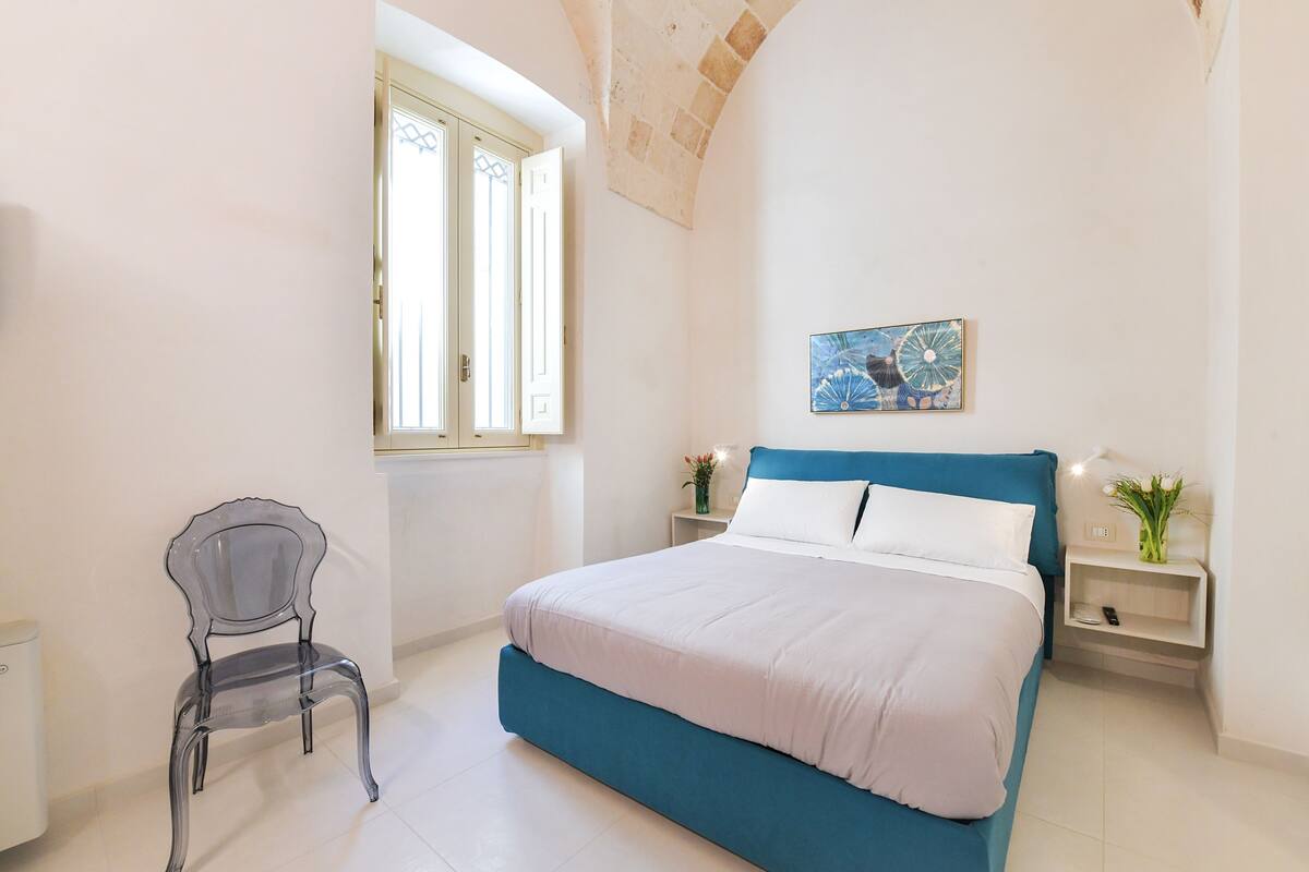 Un dormitorio con cama, una silla, un ventanal y una mesita de noche.