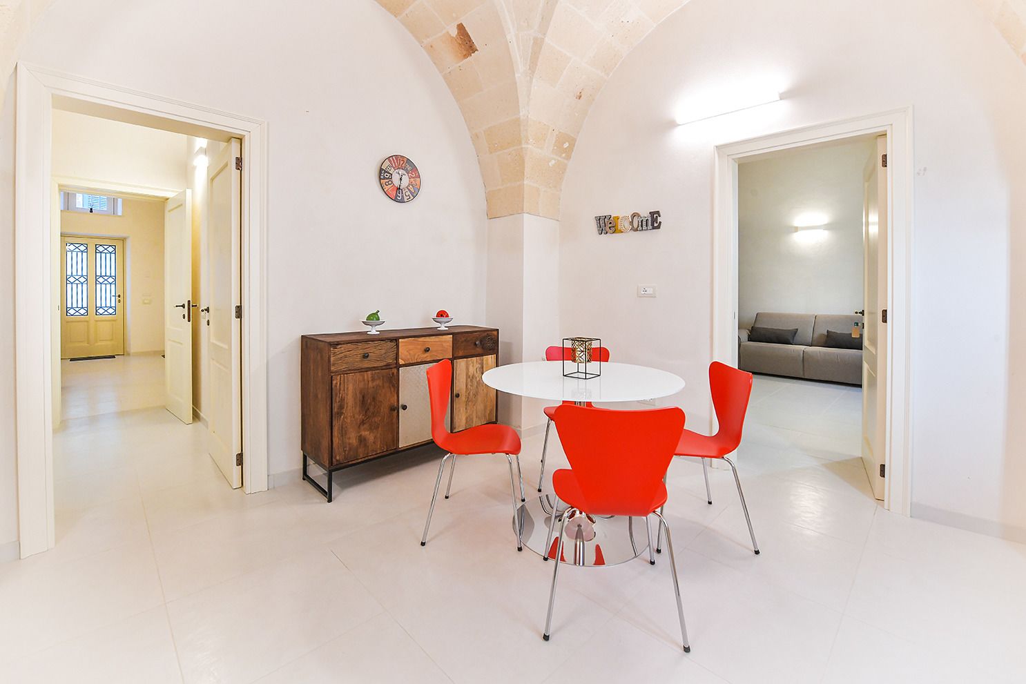 Un comedor con una mesa redonda, sillas rojas y un aparador de madera con un reloj y objetos decorativos.