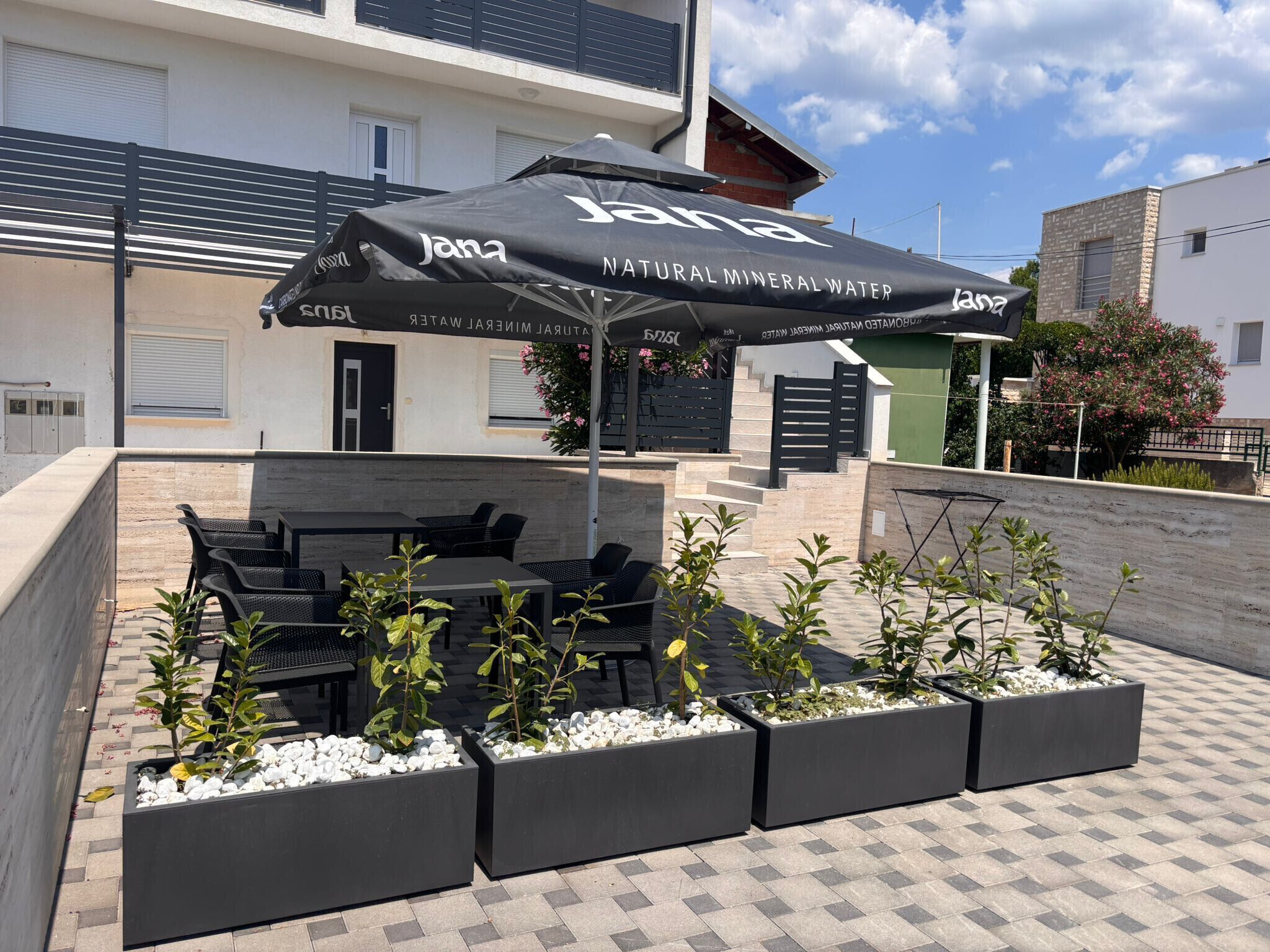 An outdoor seating area with tables and chairs, a covered umbrella, and planters with small trees.