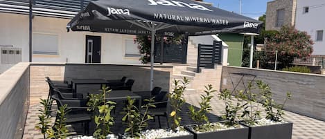 An outdoor seating area with tables and chairs, a covered umbrella, and planters with small trees.