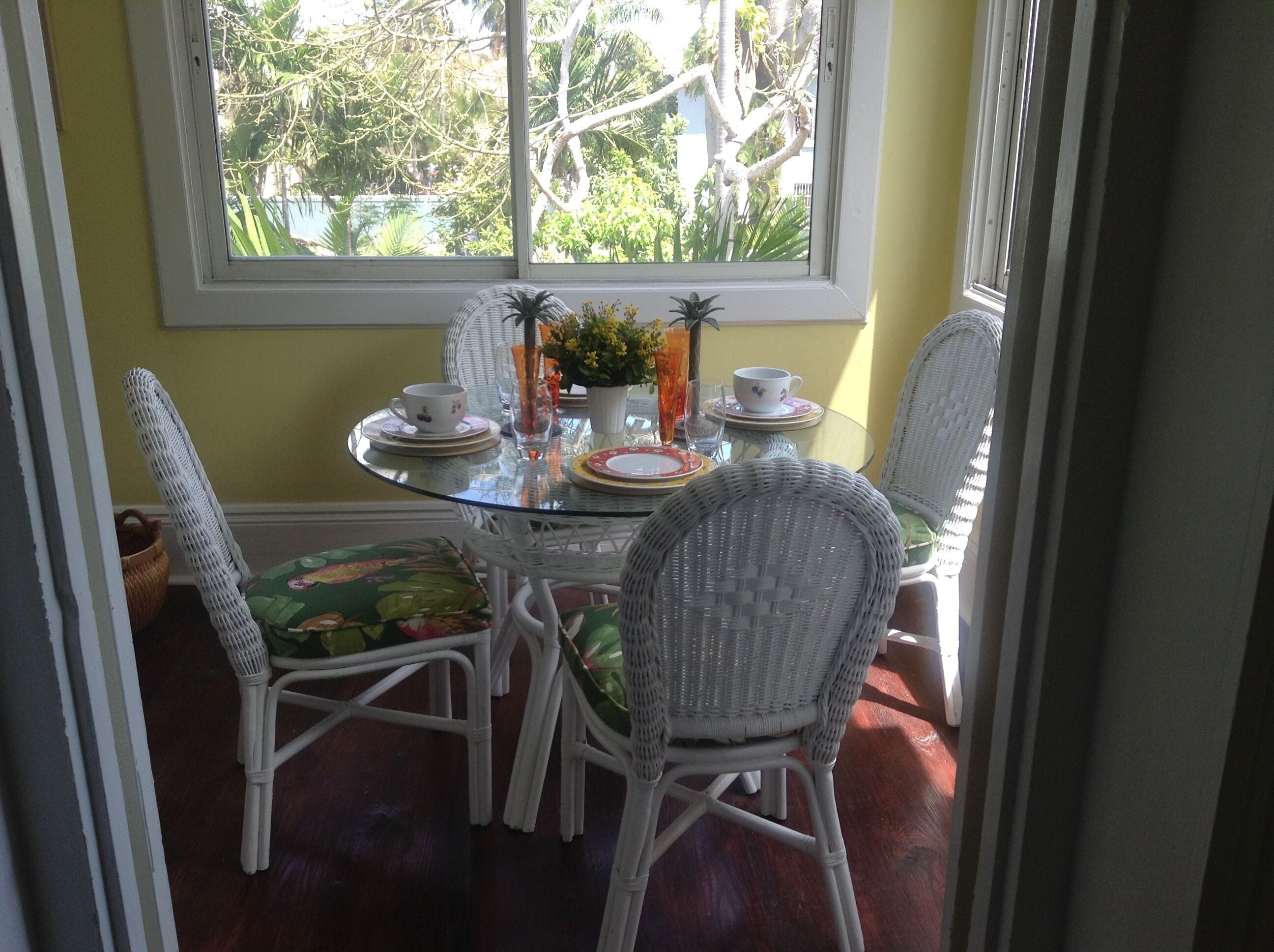A dining area with a round glass table, white wicker chairs, and a view of trees through a window.