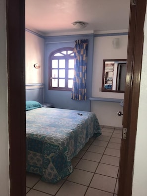 Ein Schlafzimmer mit einem Bett, einem Fenster mit Vorhängen, einem Spiegel und einer Tür.