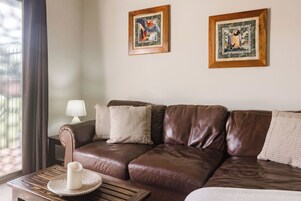 Un sofá de cuero marrón con dos cuadros enmarcados encima, una lámpara en una mesita auxiliar y una ventana con cortinas.