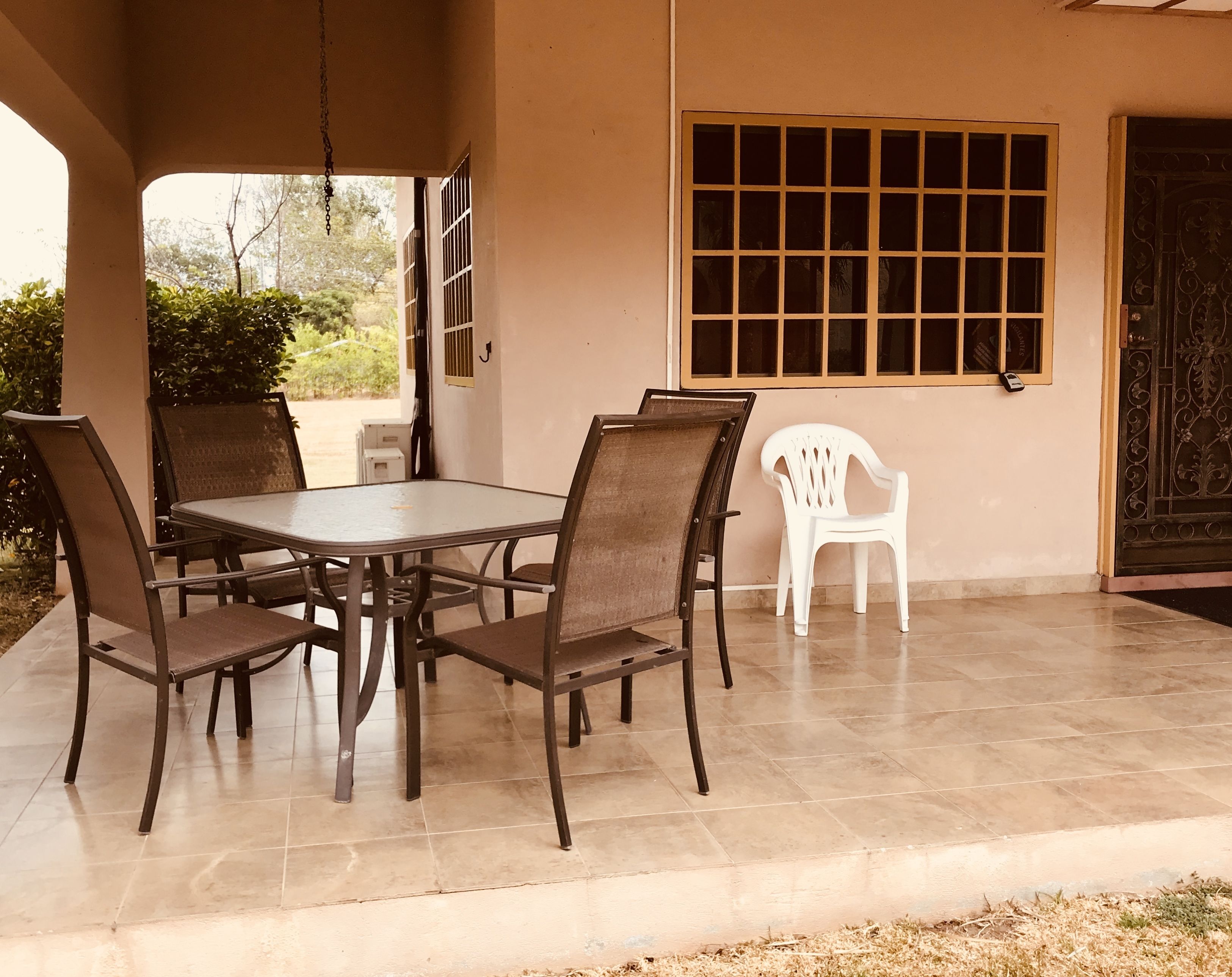 Un patio cubierto con una mesa redonda y seis sillas, una silla de plástico blanco y una ventana con barrotes.