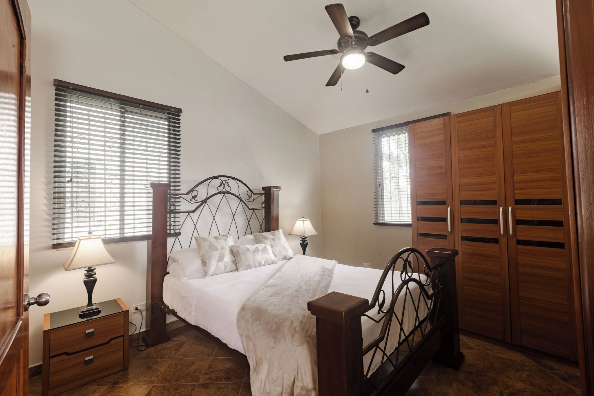 Un dormitorio con cama, armario de madera, ventilador de techo, lámpara de noche y persianas.