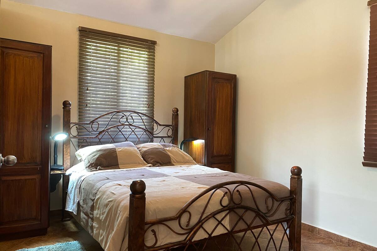 A bedroom with a wooden bed, a nightstand, a lamp, a wardrobe, and a window with blinds.