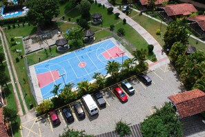 An aerial view of a sports court with a parking area, surrounded by greenery and buildings with red-tiled roofs. - Vale do Sonho Hotel & Eventos (Guararema)