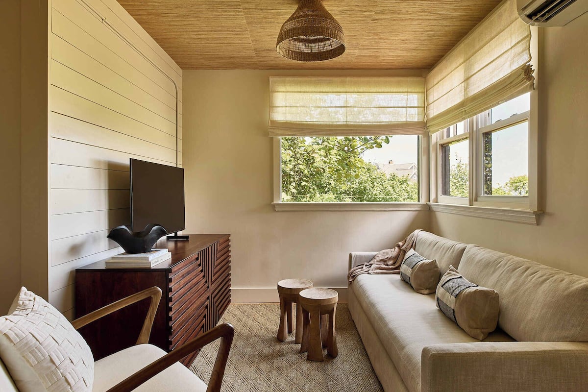 Una sala de estar acogedora con techo de madera, un televisor de pantalla plana sobre un mueble de madera, un sofá beige con cojines y un ventanal con vistas a zonas verdes.