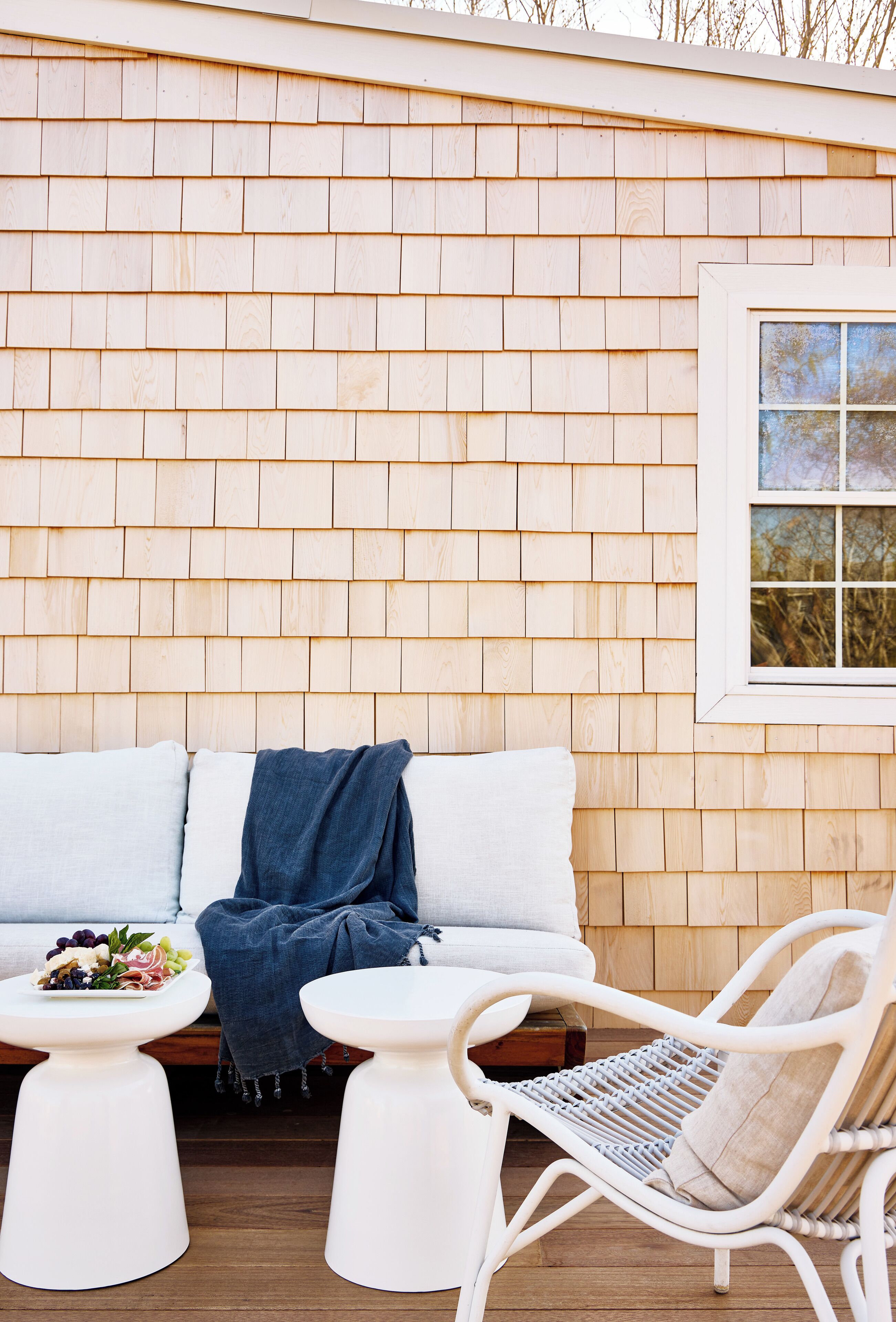A wooden exterior wall with a window, a white sofa, two white tables, and a wicker chair.