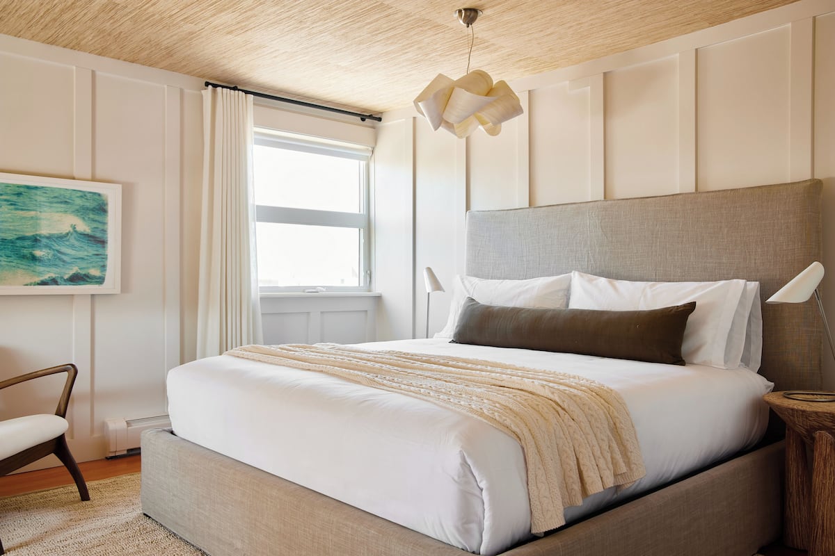 Un dormitorio con una cama grande, cabecera, mesita de noche, una silla, un cuadro en la pared y una ventana con cortinas.