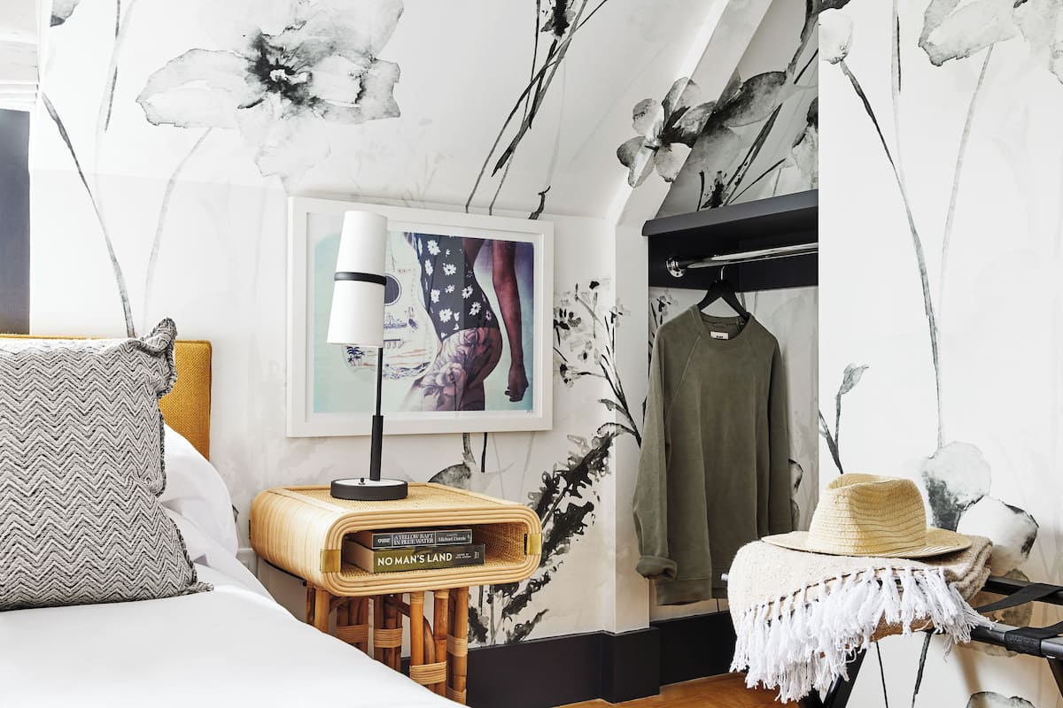 Un dormitorio con cama, una mesita de noche con libros, una lámpara, un papel tapiz floral y ropa colgada.