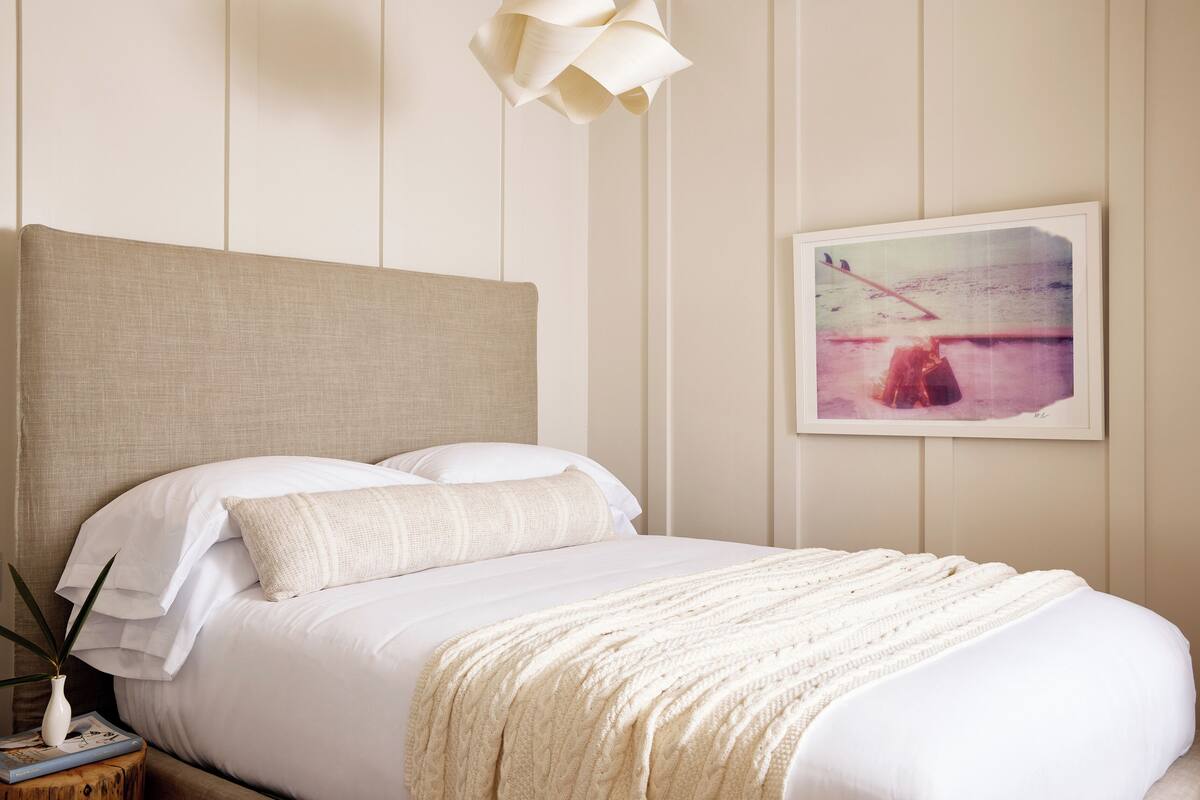 Una cama bien tendida con sábanas blancas y un plaid color crema. Un cuadro enmarcado cuelga en la pared sobre la cama.