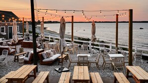 Ett utomhusområde med sittplatser, bestående av träbord och stolar, ljusslingor och parasoller, med utsikt över stranden.