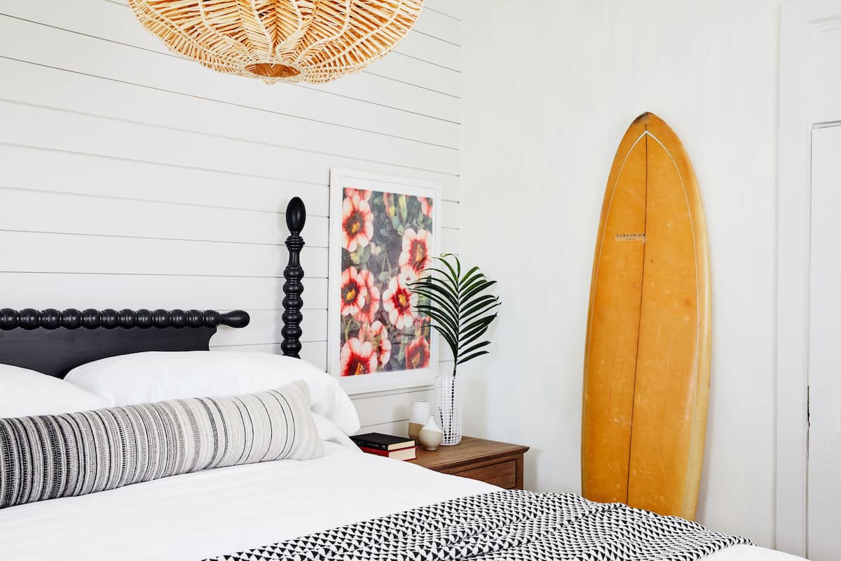 Un dormitorio con cama, mesita de noche, una tabla de surf y un cuadro floral en la pared.