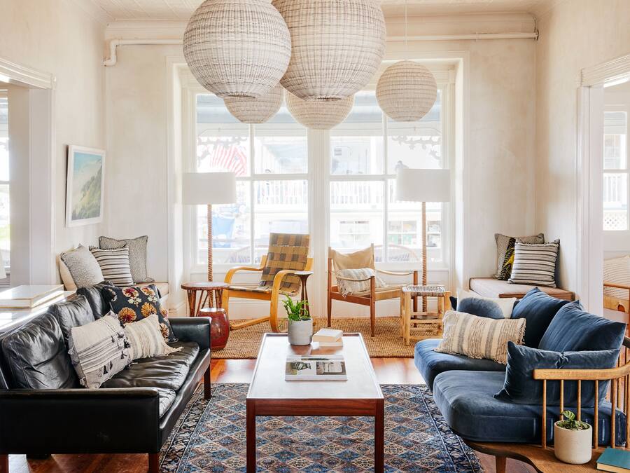 A spacious living room with a black leather sofa, a blue sofa, and a wooden coffee table. There are wicker lamps hanging from the ceiling and a large window providing natural light.