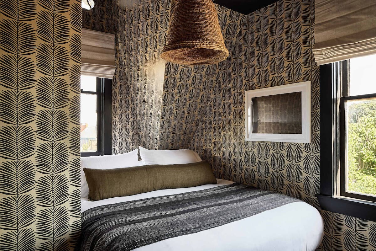 Un dormitorio con cama, lámpara colgante, ventana y un cuadro en la pared.