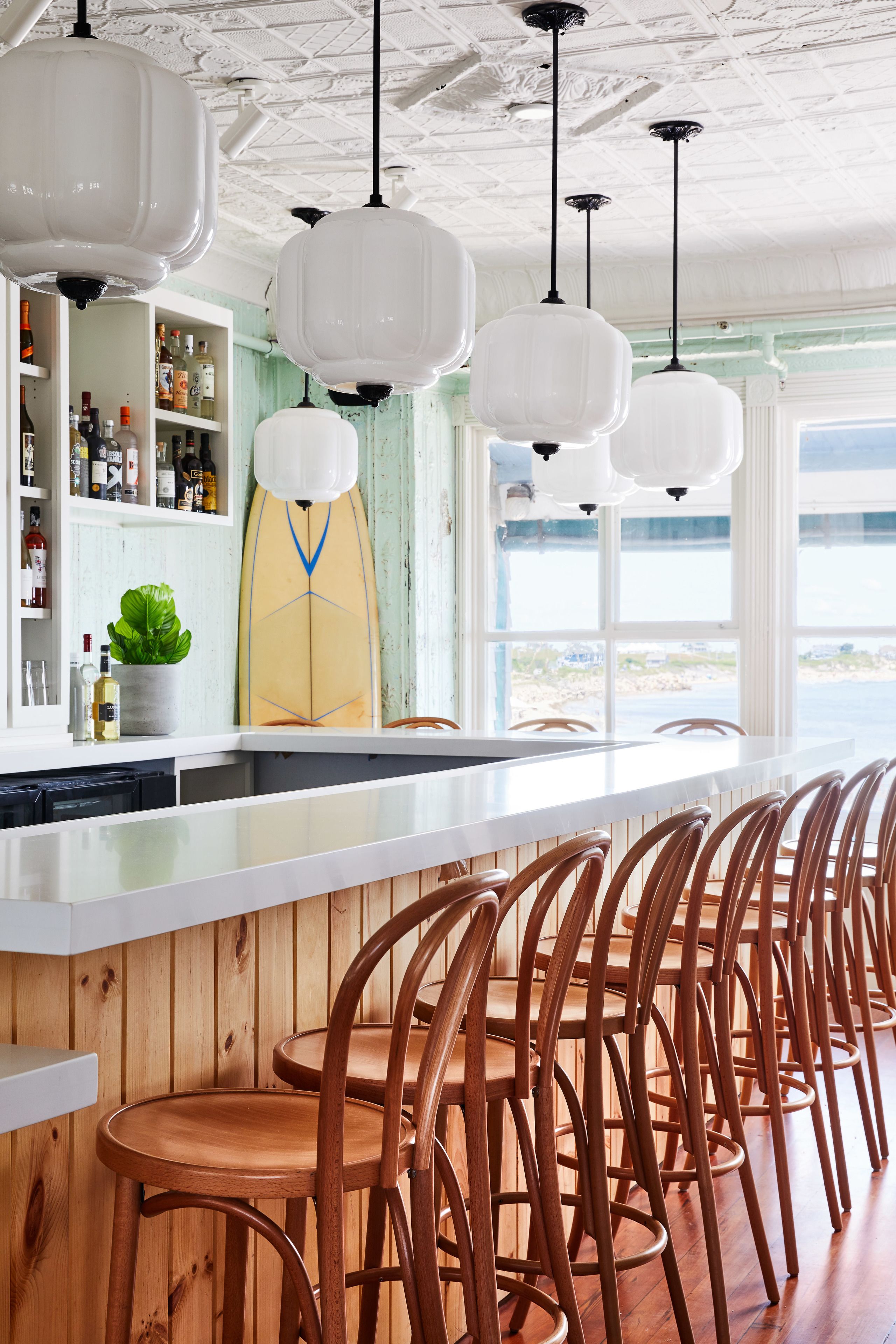 A bar area with wooden chairs, white pendant lights, and a surfboard decoration.