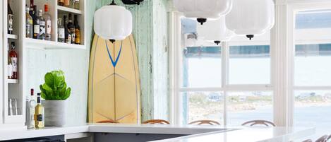 A bar area with wooden chairs, white pendant lights, and a surfboard decoration.