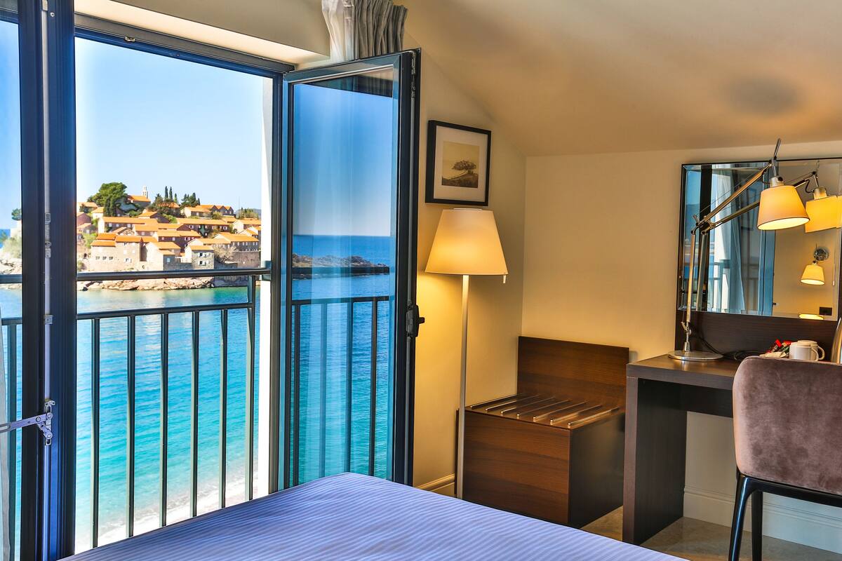 Une chambre d’hôtel dotée d’une grande fenêtre donnant sur la mer et les bâtiments, d’un lit, d’un bureau avec une chaise et d’une lampe.