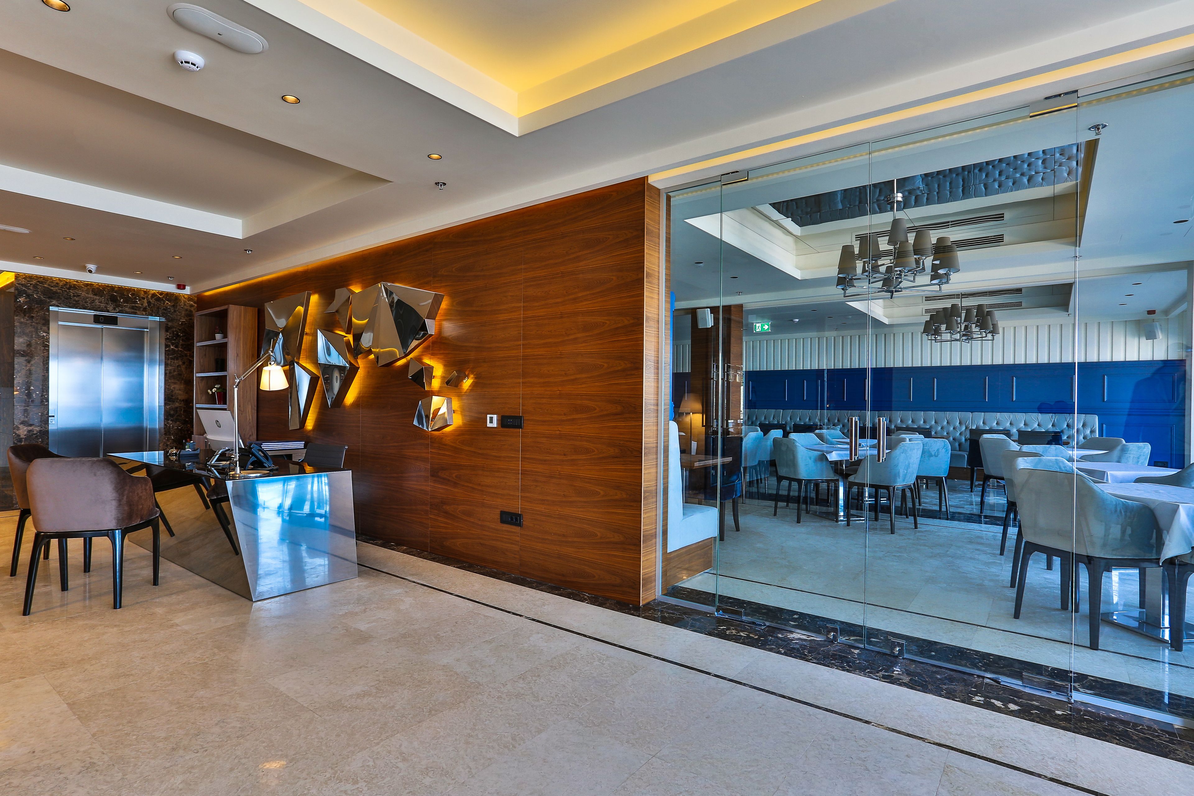 Un moderno vestíbulo de hotel con mostrador de recepción, zona de estar y vistas al área de comedor a través de un panel de vidrio.