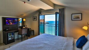 Ein Hotelzimmer mit Bett, Schreibtisch mit Computer, Minikühlschrank, Stuhl und einem Balkon mit Meerblick.