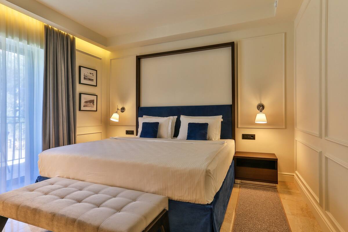 Une chambre d’hôtel bien aménagée, avec un grand lit, un banc, deux tables de chevet et une vue sur l’extérieur.