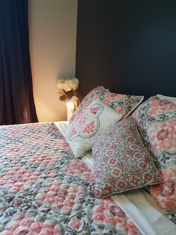 Una cama con ropa de cama y almohadas de estampado floral, una mesita de noche con un florero y una pared de color oscuro.