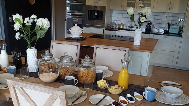 Et moderne køkken med et træbord dækket til morgenmad, inklusive kaffemaskine, syltetøjsglas og en vase med blomster.