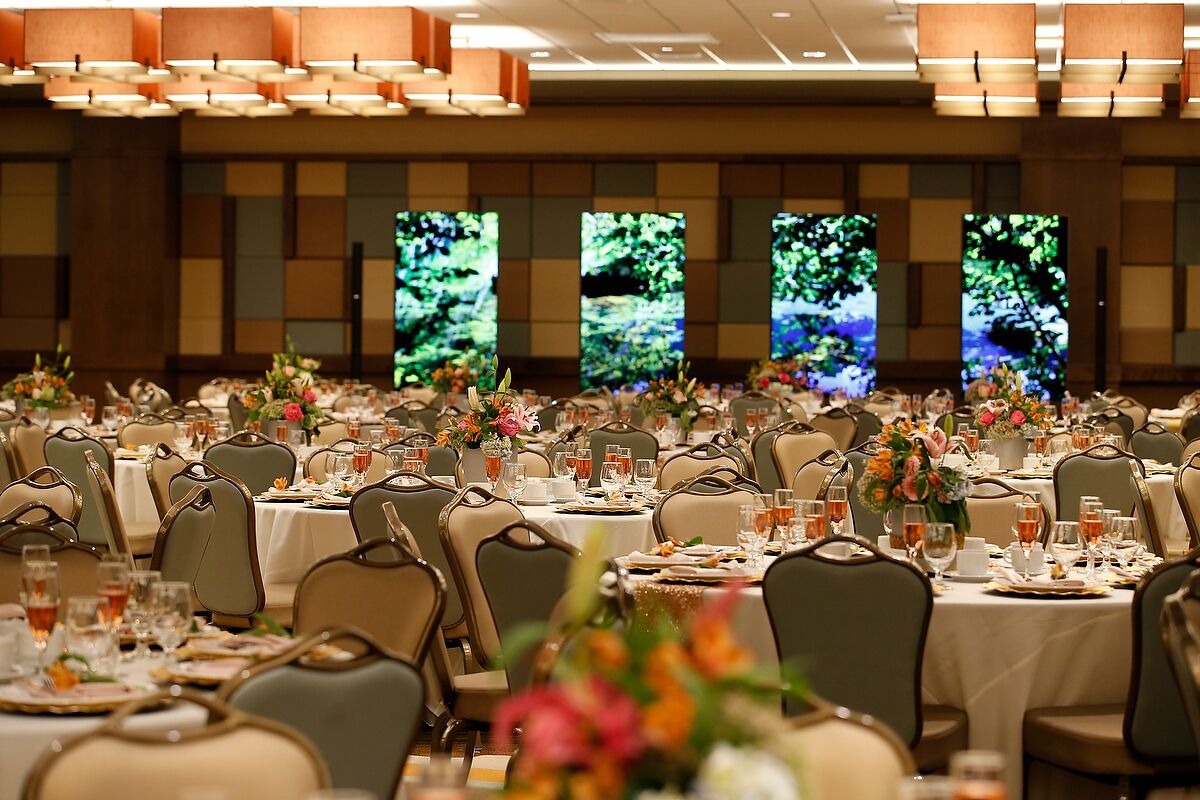Un salón de banquetes con varias mesas redondas, cada una dispuesta con sillas, vasos y centros de mesa florales.
