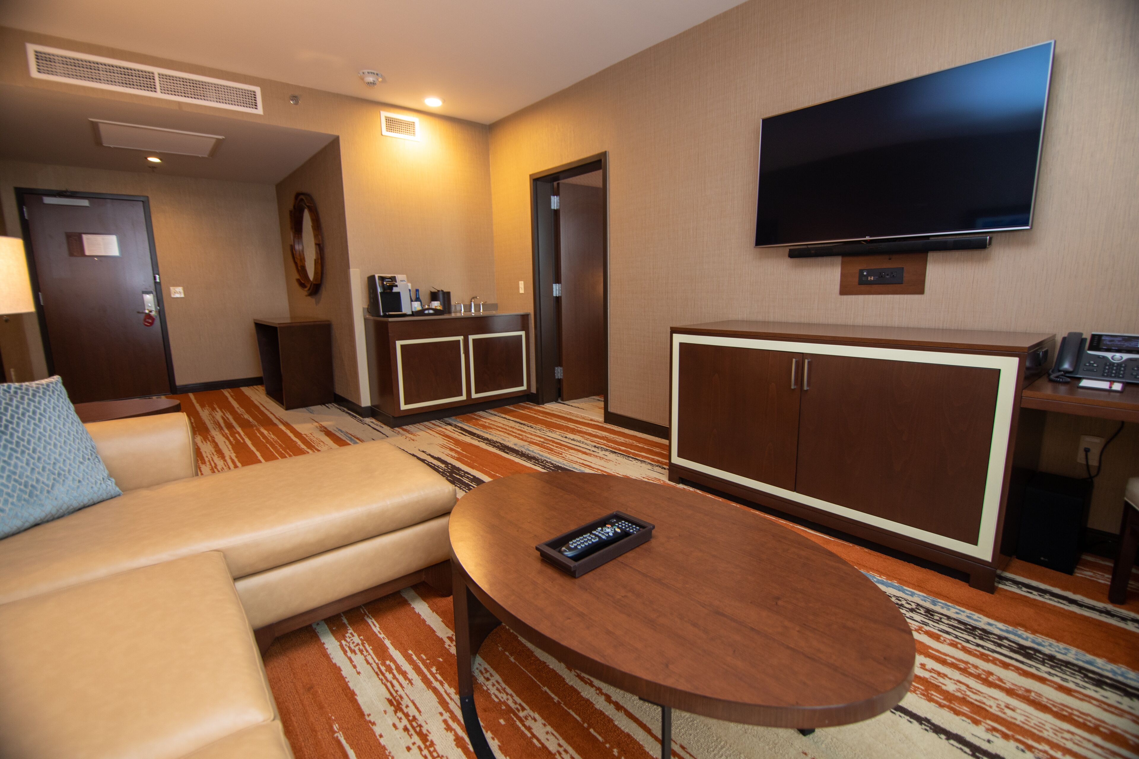 Habitación de hotel con sofá, una mesita redonda, televisor montado en la pared, minibar y escritorio con teléfono.