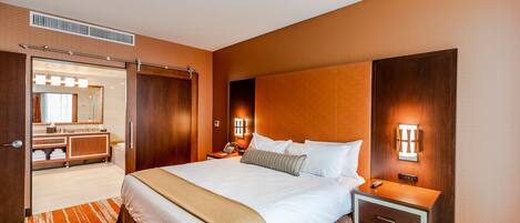A hotel room with a large bed, a bedside table with a lamp, a nightstand, a wooden headboard, and a bathroom visible through an open door.
