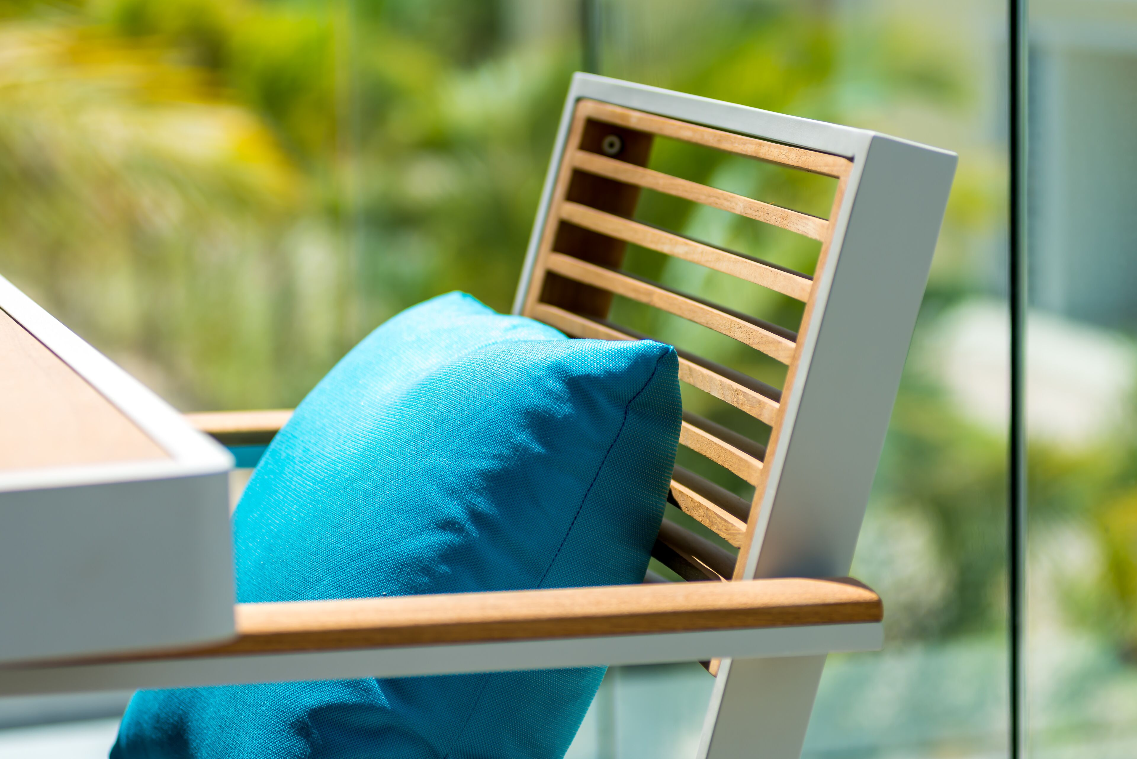 A chair with a wooden backrest and a blue cushion, positioned near a window with a blurred green background.