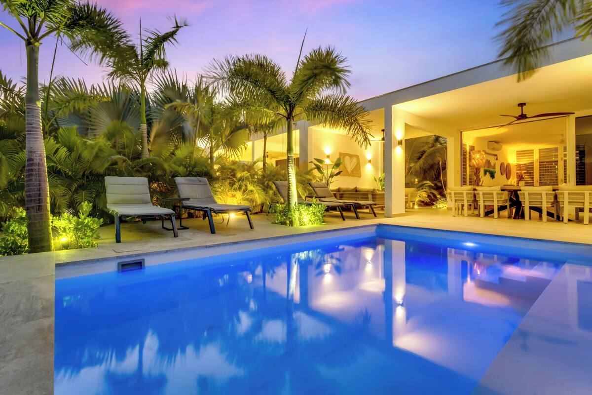Uma piscina com espreguiçadeiras, palmeiras e um espaço interno bem iluminado.