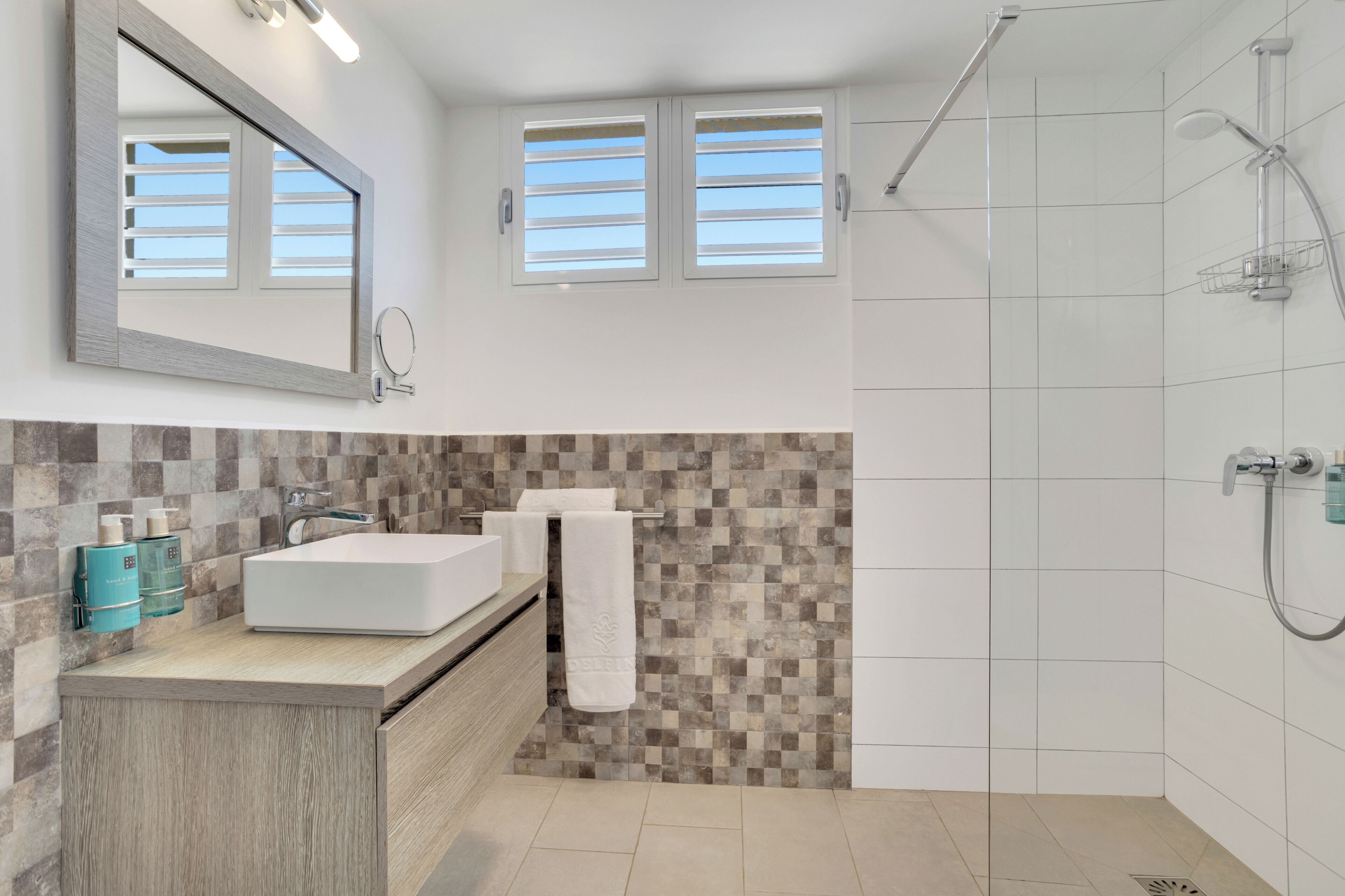 A modern bathroom with a large mirror, two windows, a shower, and a vanity with a sink and towel rack.