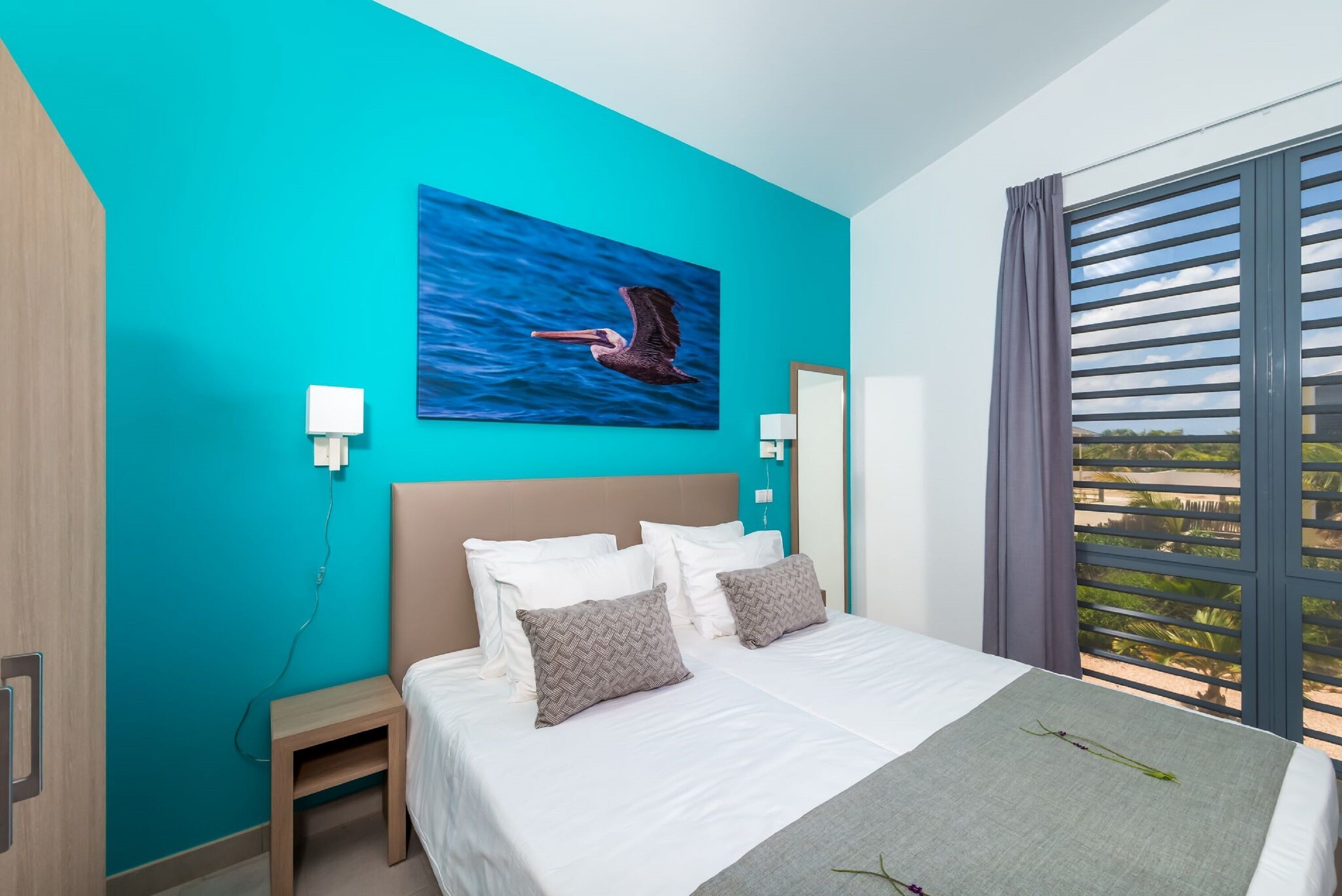 A modern hotel room with a large bed, teal walls, a painting of a pelican, and a view through the window.