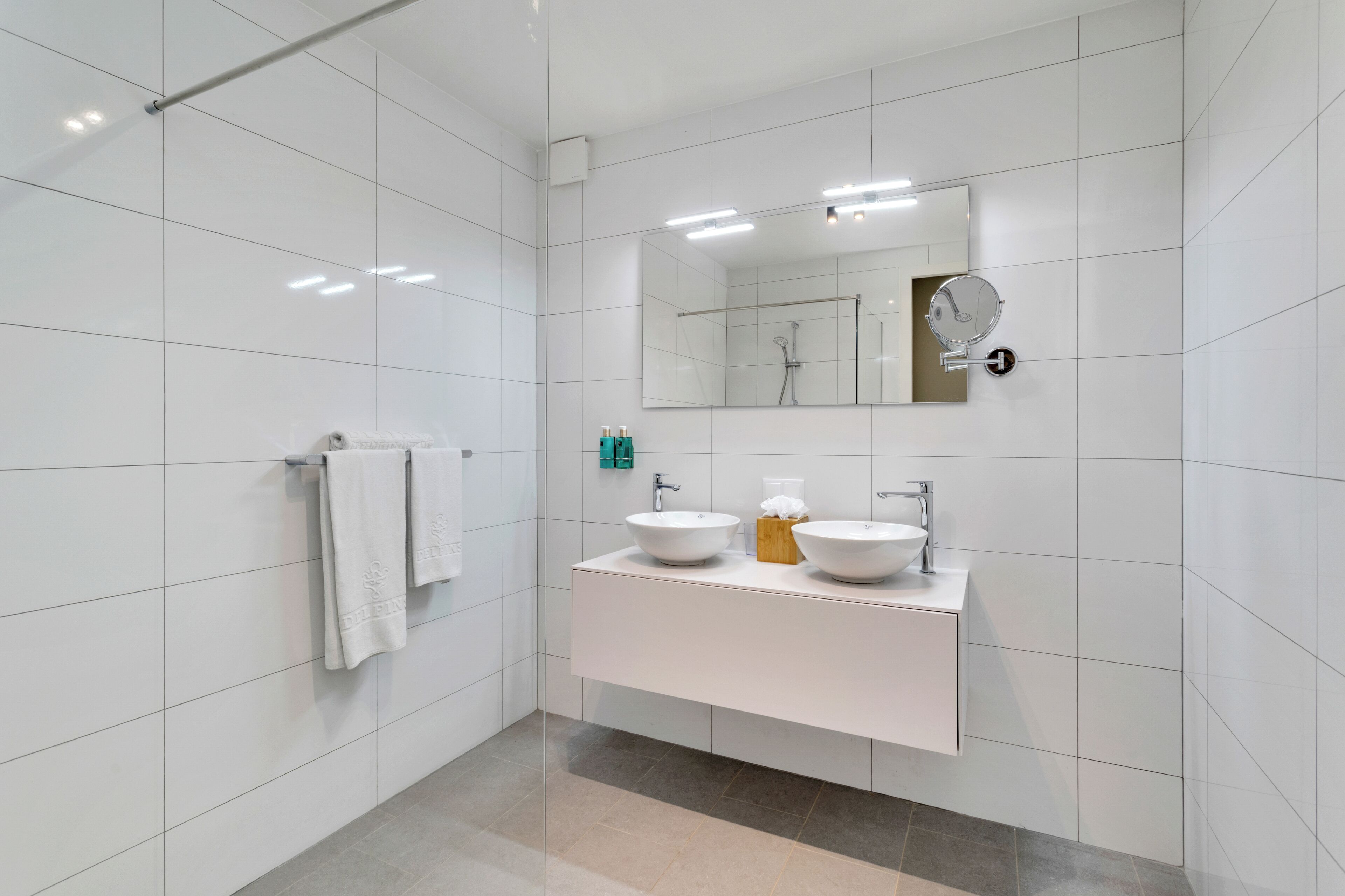 A modern bathroom with white tiled walls, a large mirror, two sinks, and a shower.