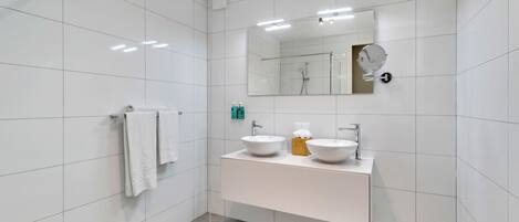 A modern bathroom with white tiled walls, a large mirror, two sinks, and a shower.