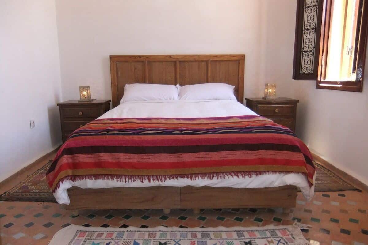 Uma cama com cabeceira de madeira, duas mesas de cabeceira com luminárias e um tapete estampado sobre um piso de azulejos.