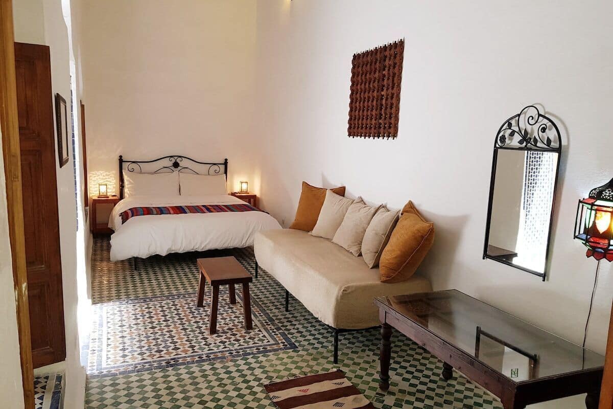 Quarto com cama, sofá, uma pequena mesa, espelho e piso estampado.