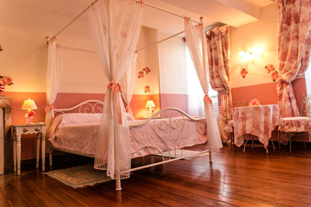 A four-poster bed with a canopy, a bedside table with a lamp, a chair with a table, and a wall-mounted light fixture.