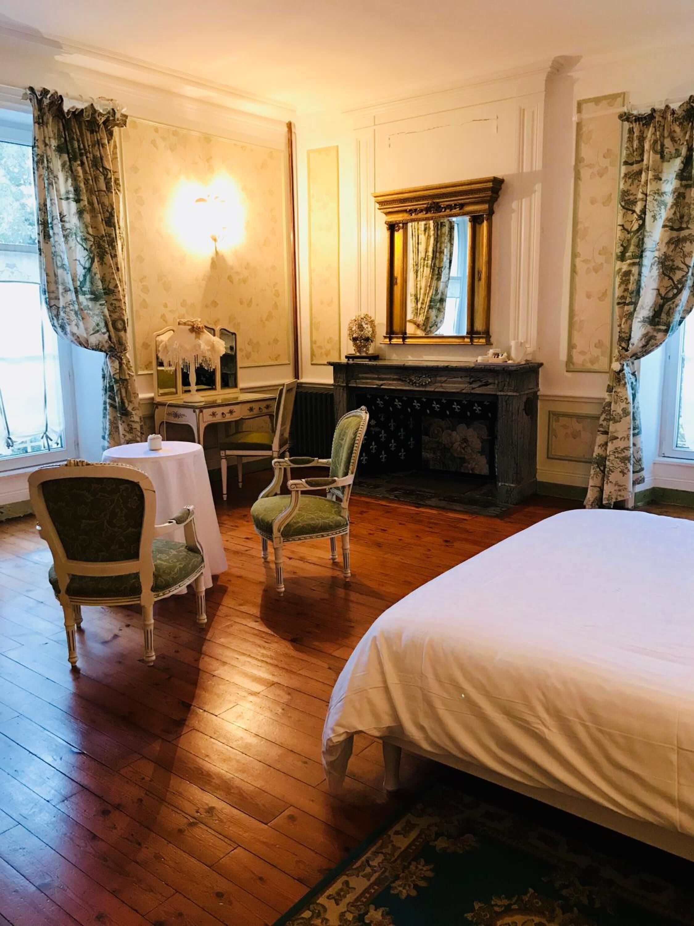 Une chambre de style traditionnel, dotée d’un grand lit, de deux fauteuils, d’une petite table, d’une cheminée et d’un miroir.