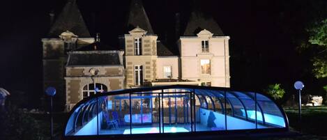 Un'ampia struttura in vetro illuminata con una piscina di fronte a un edificio storico di notte.