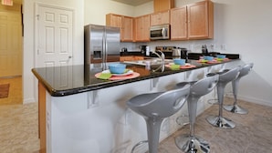 A kitchen with wooden cabinets, a stainless steel refrigerator, a microwave, a sink, and three bar stools.