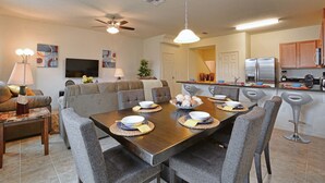 A modern kitchen with a dining area, featuring a large wooden table, grey upholstered chairs, and a ceiling fan.