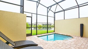 An indoor swimming pool with tiled flooring and lounge chairs, surrounded by glass walls offering a view of buildings and greenery.